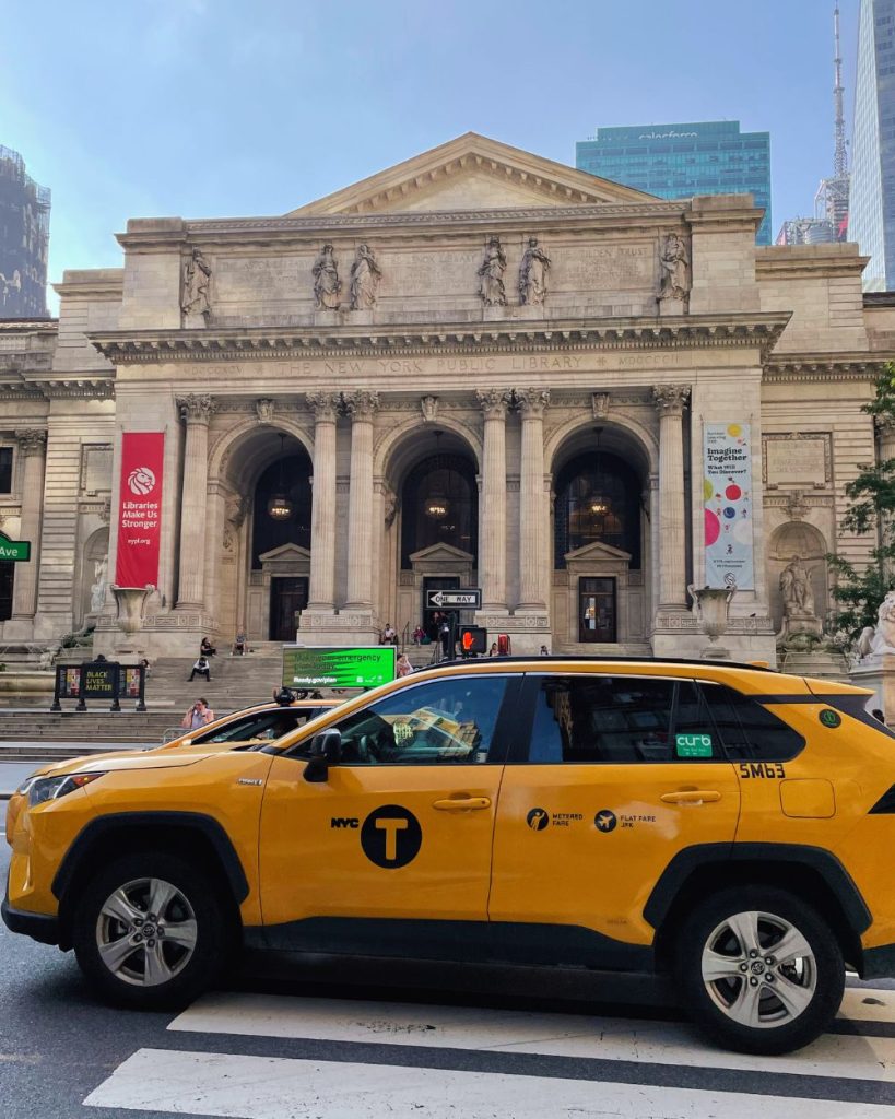 Fachada principal de la Biblioteca Pública de Nueva York (NYPL) con las escaleras de mármol y un taxi amarillo circulando por la Quinta Avenida.