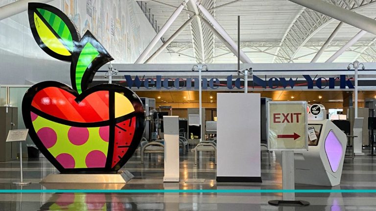 Cartel de Welcome to NYC con una manzana colorida en la terminal del Aeropuerto JFK, portada de la guía de traslado a Manhattan.