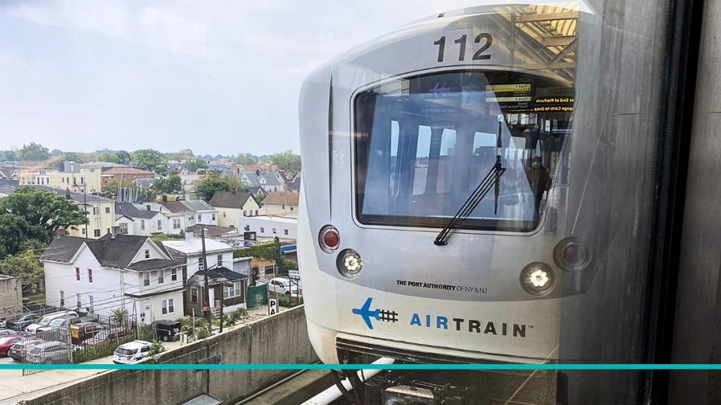 Vista del AirTrain de JFK circulando por las vías elevadas sobre el barrio de Queens, con el horizonte de la ciudad de fondo.