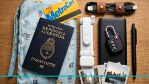 Composición de artículos indispensables para el viaje: pasaporte argentino, cargador portátil, candado de maleta, trípode compacto y la MetroCard de Nueva York sobre una superficie neutra.