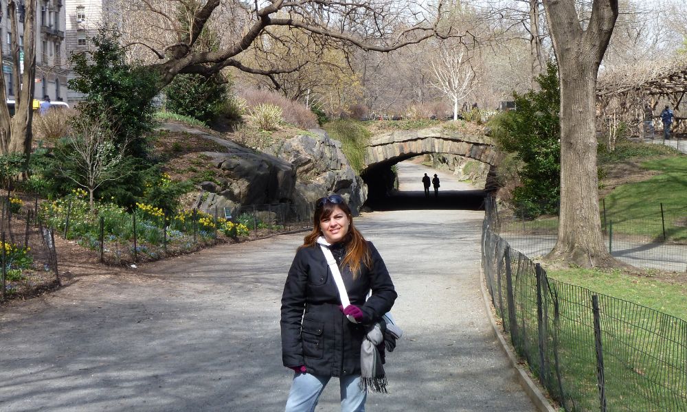 Una mujer posando en un sendero de Central Park durante el comienzo de la primavera, rodeada de árboles grises sin hojas antes del florecimiento.
